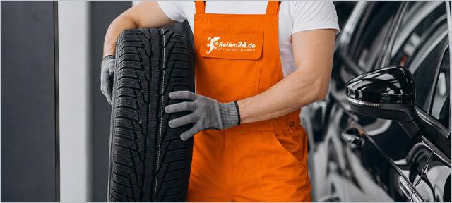 Reifen24-Mitarbeiter in orangefarbener Arbeitskleidung und grauen Handschuhen hält einen Winterreifen neben einem schwarzen Auto.